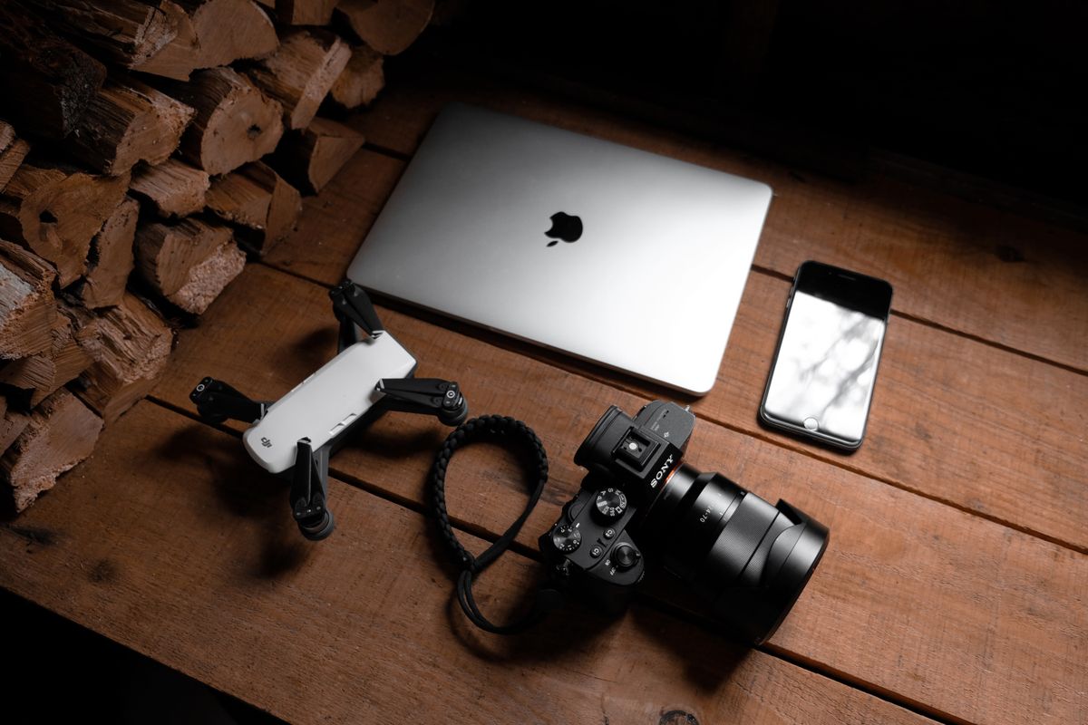 A flat lay of modern technology devices including a laptop, camera, drone, and smartphone on a wooden table.