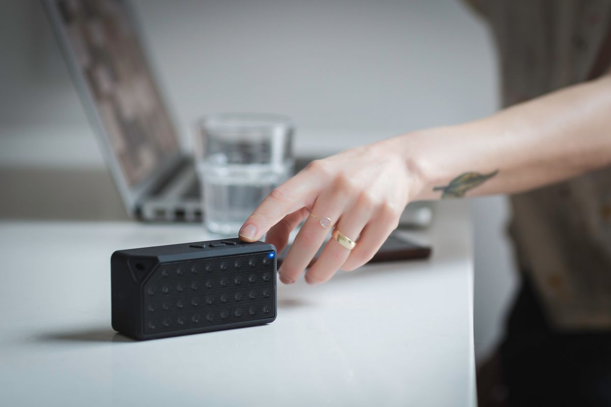 Close-up of a person adjusting a portable Bluetooth speaker on a desk indoors, with a laptop in the background.