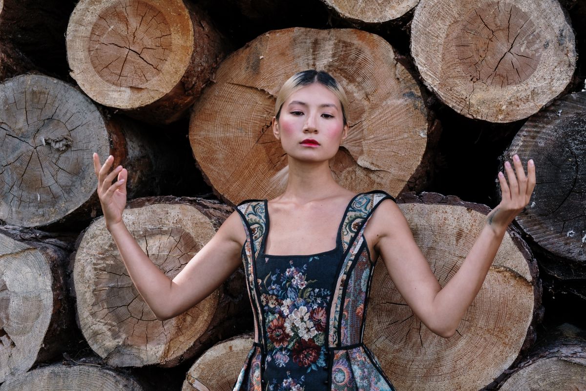 A stylish model in a floral dress poses gracefully in front of stacked wooden logs.