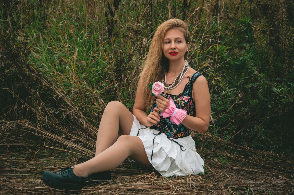 A woman in a floral dress sits in a grassy field holding a pink rose, exuding a natural elegance.