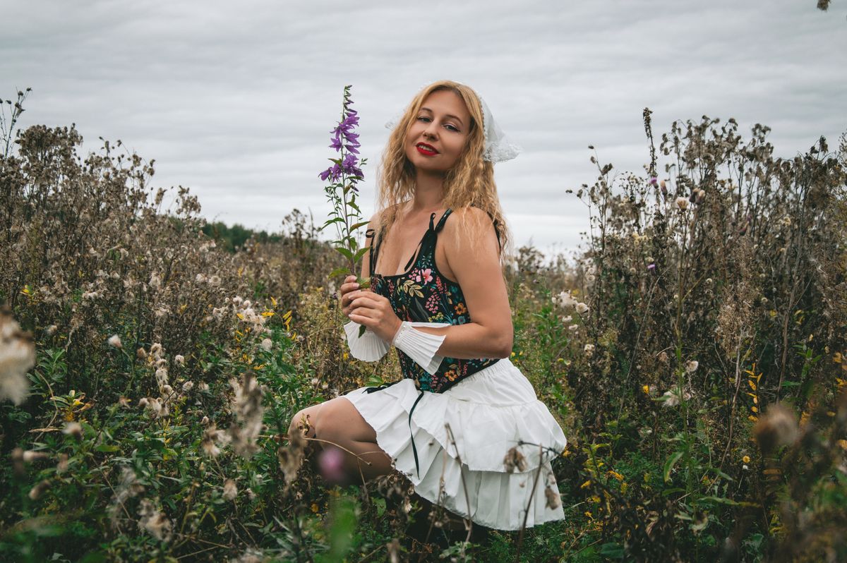 A woman in a floral dress holds purple flowers while kneeling in a field under a cloudy sky.