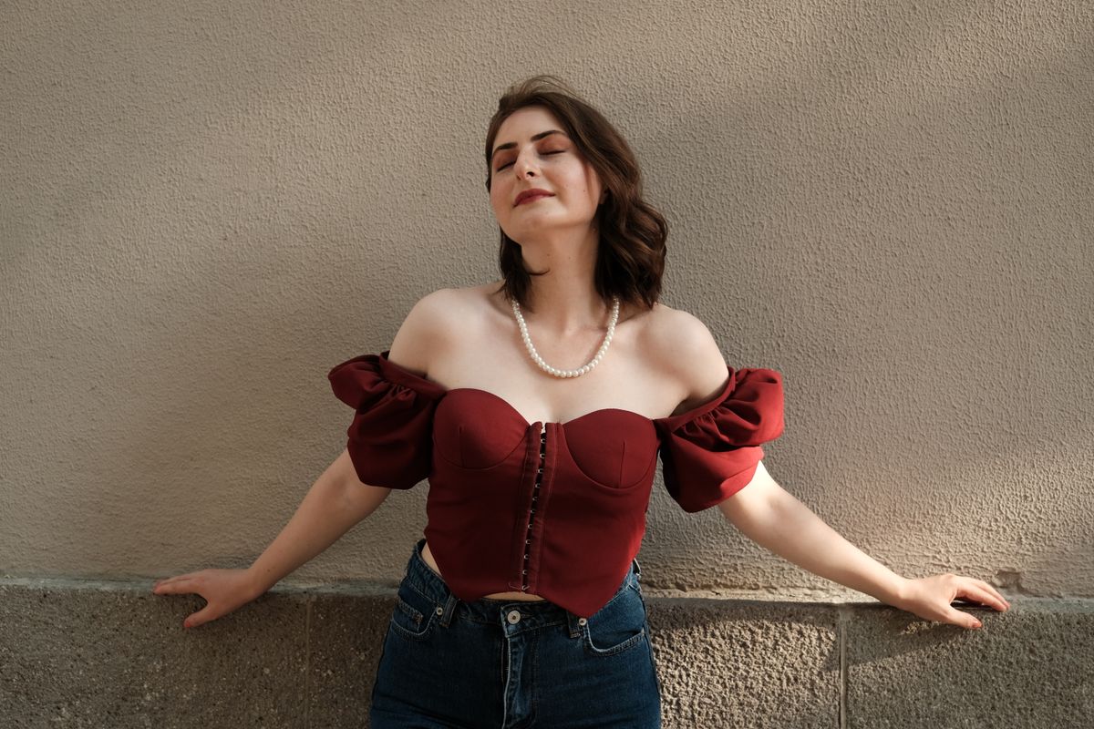 Portrait of a woman with brown hair and eyes closed, wearing a stylish red top with an off-shoulder design and pearls.