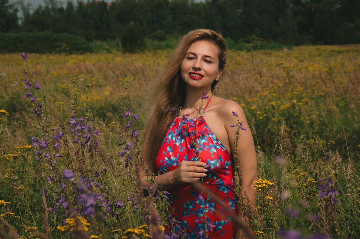 Cheerful woman in a red floral dress enjoying a walk in a vibrant wildflower field under the summer sun.