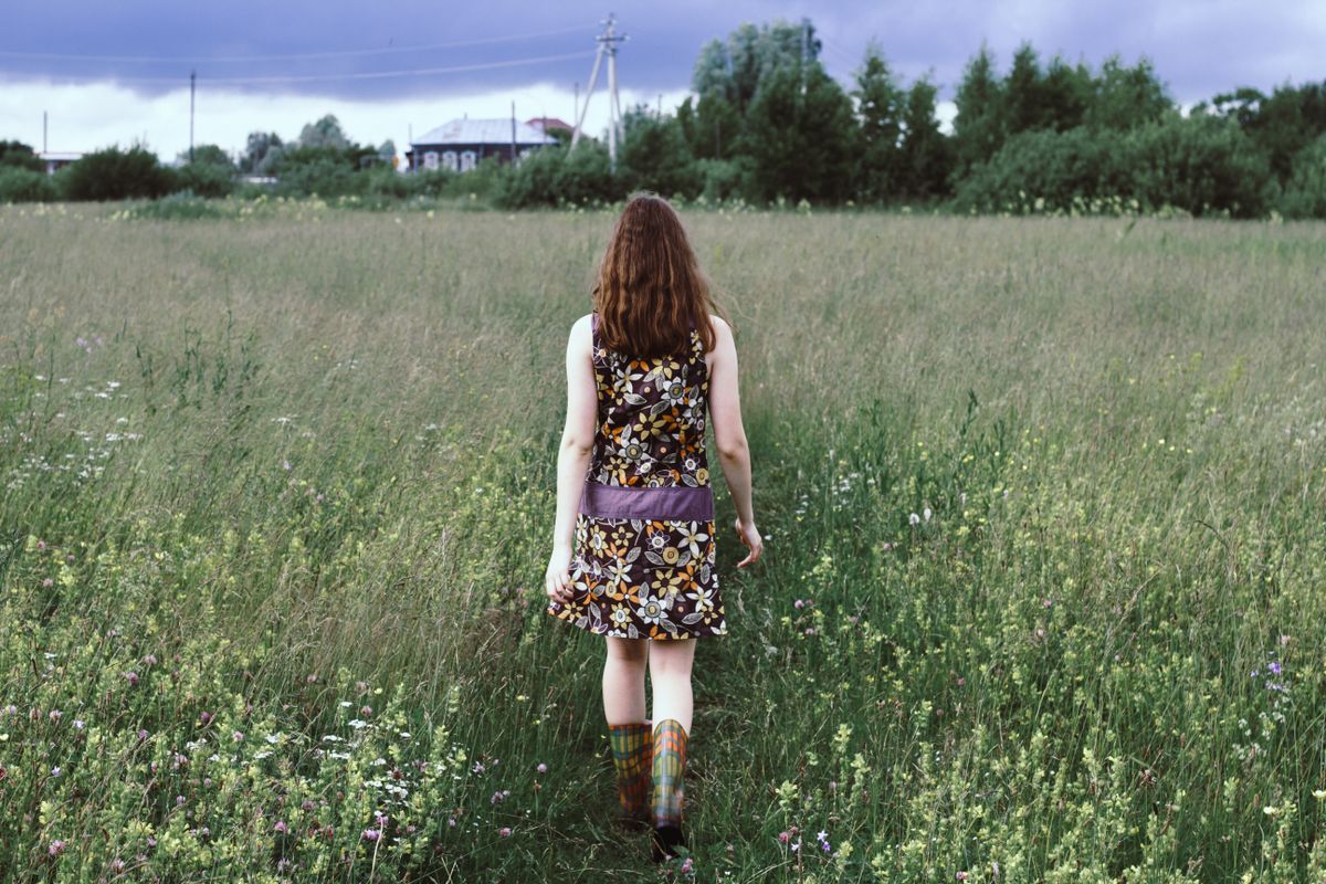 A woman walks alone through a lush grass field, showcasing a floral dress.