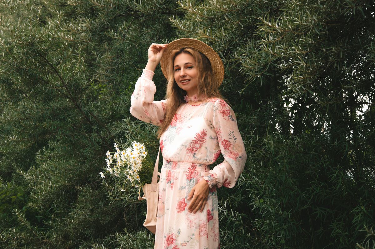 A woman wearing a floral dress and straw hat poses outdoors with green foliage.
