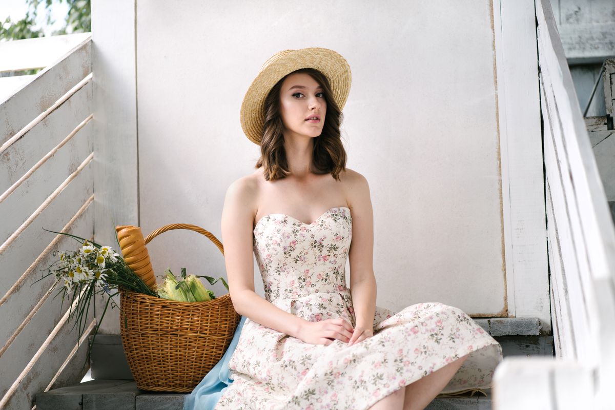 A woman in a floral dress and straw hat sits on stairs with a basket of groceries.