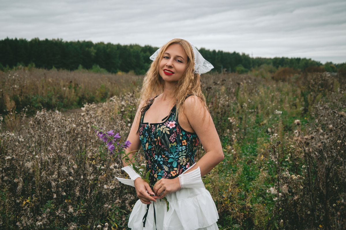 Woman in floral dress holding flowers in a lush field under cloudy sky.