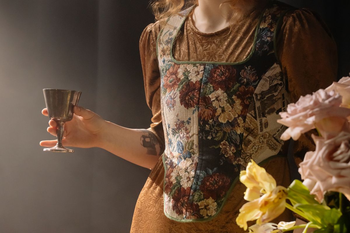 A woman in a vintage floral dress holds a wine glass next to soft pink flowers.