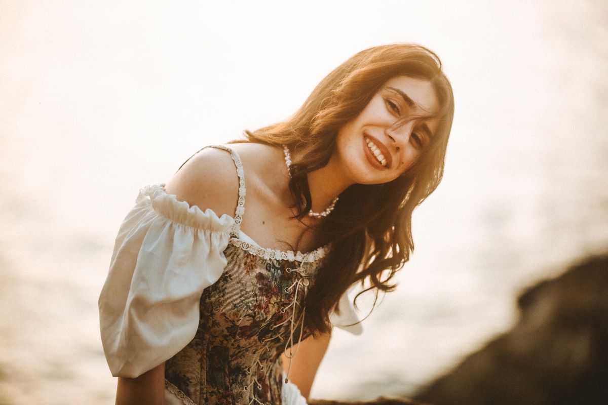 A cheerful woman in a vintage dress smiling brightly against a blurred outdoor background.