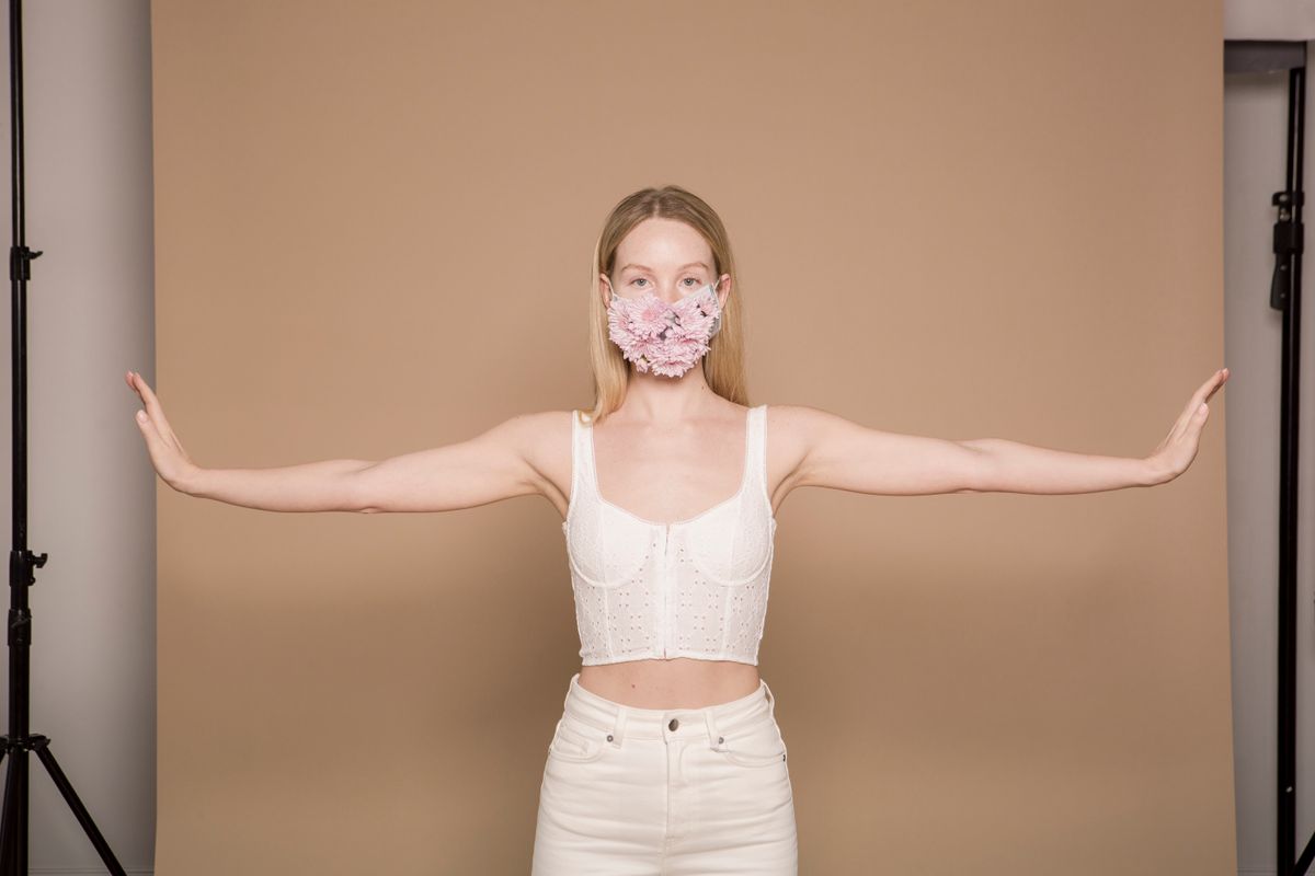 Serious female in white outfit wearing flower mask and looking at camera with arms spread in modern studio during pandemic