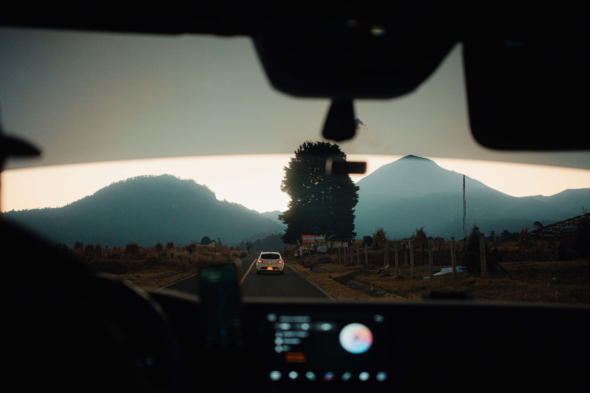 A car drives on a scenic road framed by mountains at sunset, viewed from a vehicle interior.