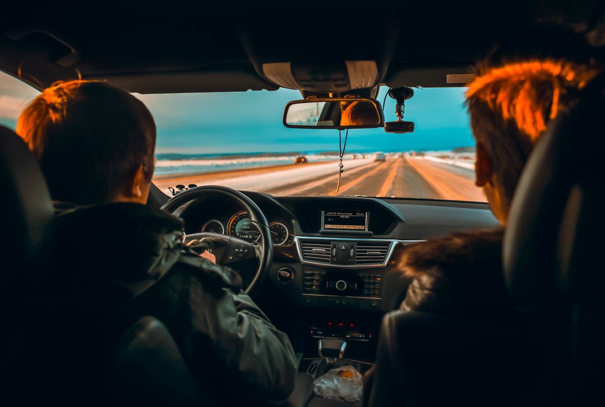 Interior view of a car driving down a snowy road at sunset with two people in the front seats.
