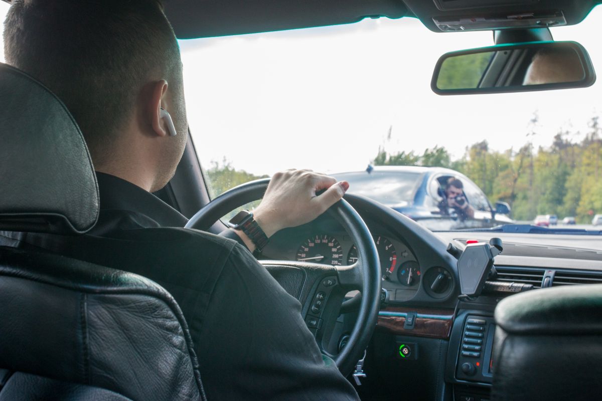 A man is driving a car seen from the back seat, focusing on the road ahead.