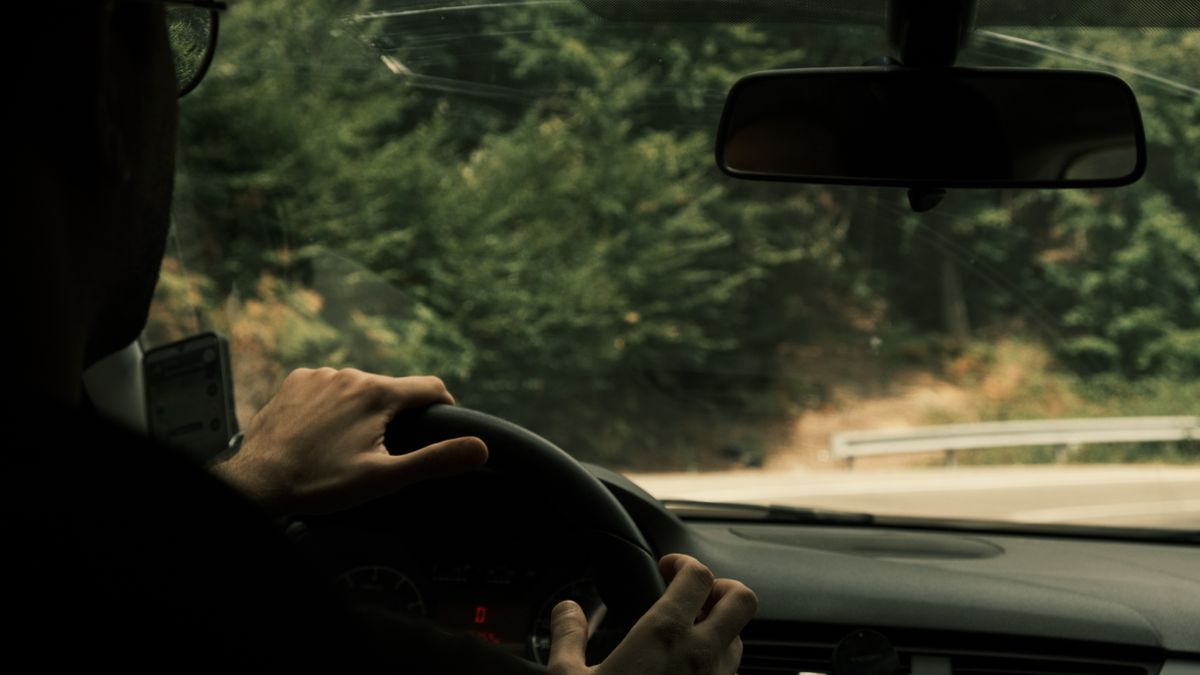 Person driving through forested road, focus on steering wheel and hand.