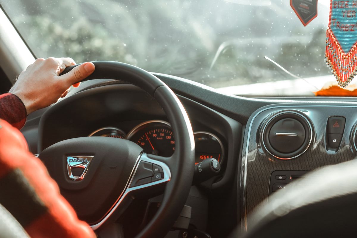 A close-up of a driver's hand on a car's steering wheel inside a vehicle with visible dashboard.
