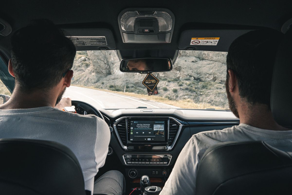 Two men driving through scenic landscape, captured from car interior showing dashboard and road.