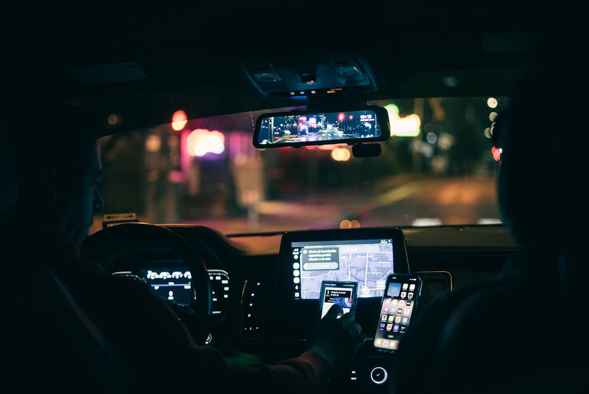 Interior view of a car at night with a driver using a smartphone for navigation through city streets.