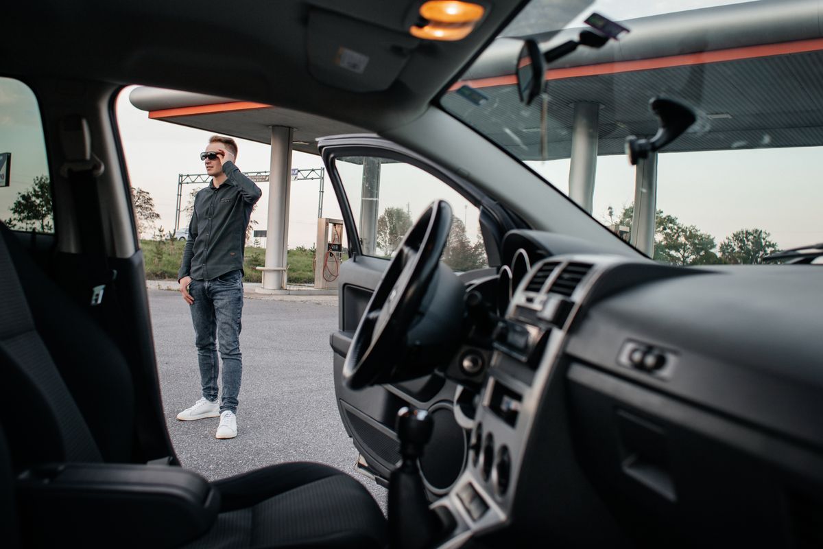 A man stands beside a car at a gas station, viewed from the car interior.