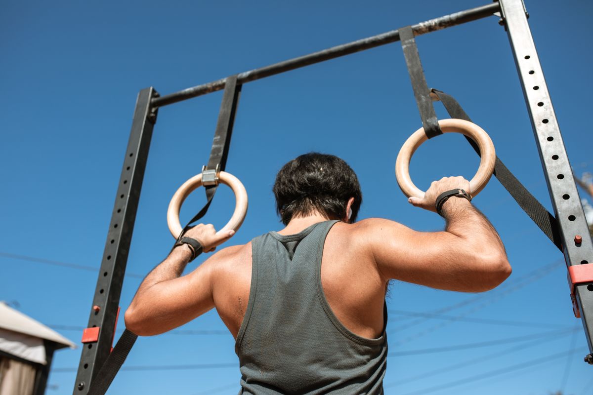 Athletic man doing a pull-up exercise on gymnastic rings outside under a clear sky.