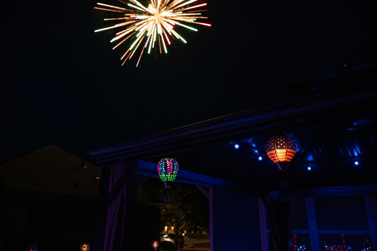 Colorful fireworks burst in the night sky above a patio with festive lanterns and lights.