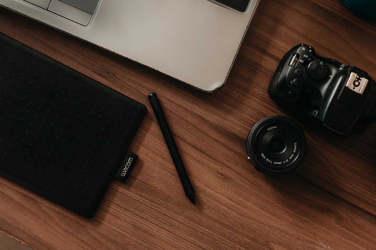 A top view of a digital workspace featuring a camera, tablet, pen, and laptop on a wooden desk.