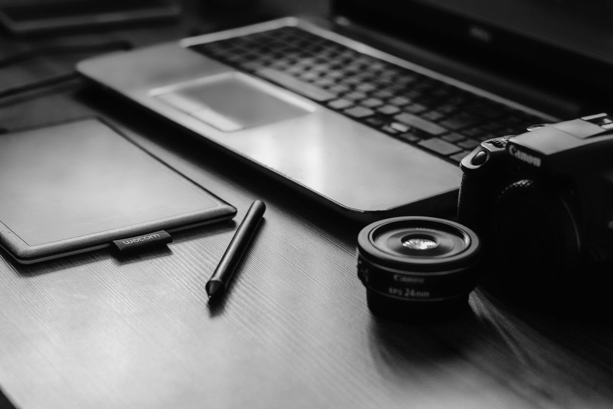 A monochromatic workspace with a camera, laptop, lens, and tablet, ideal for photographers.