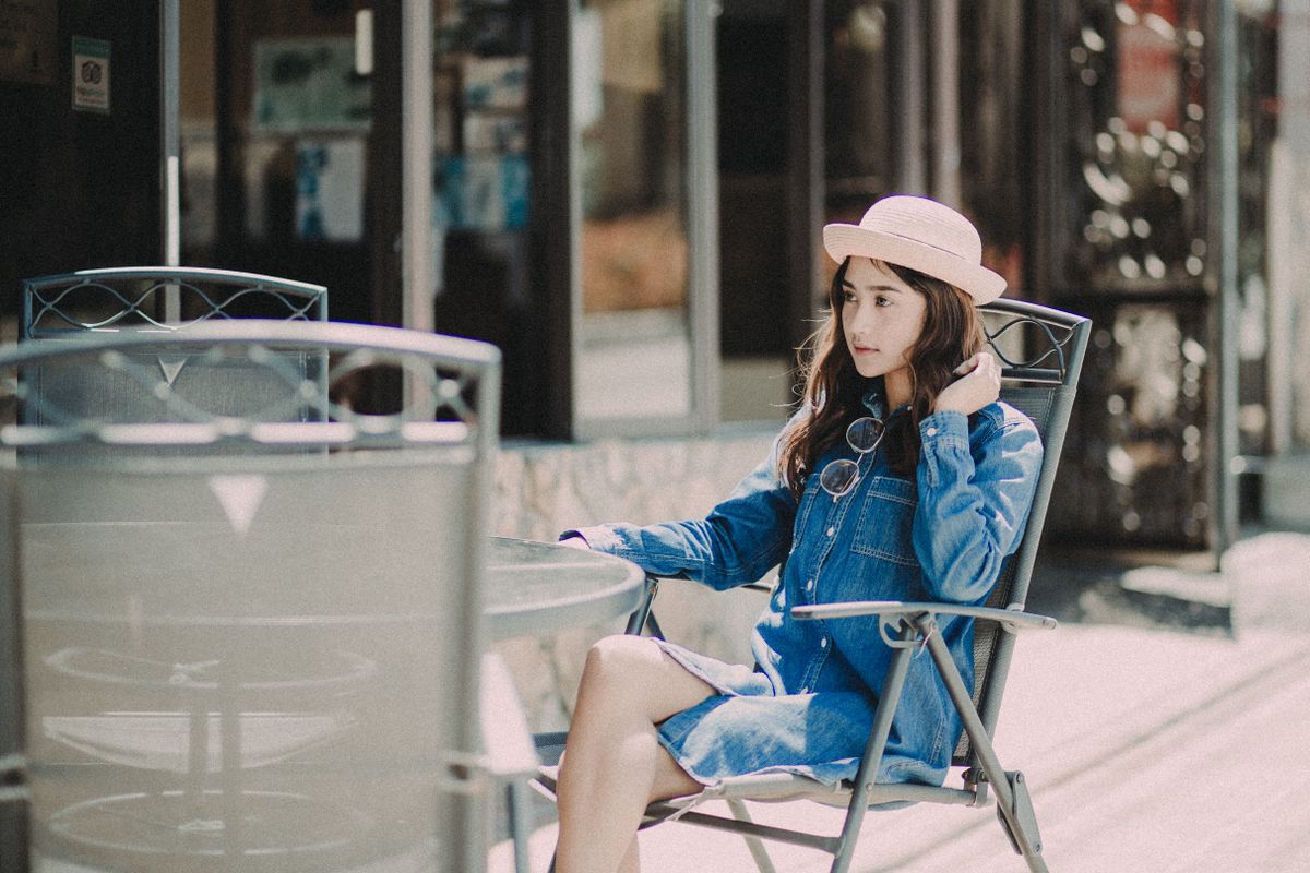 A young woman in a denim dress and hat sits outside at a cafe table on a sunny day.