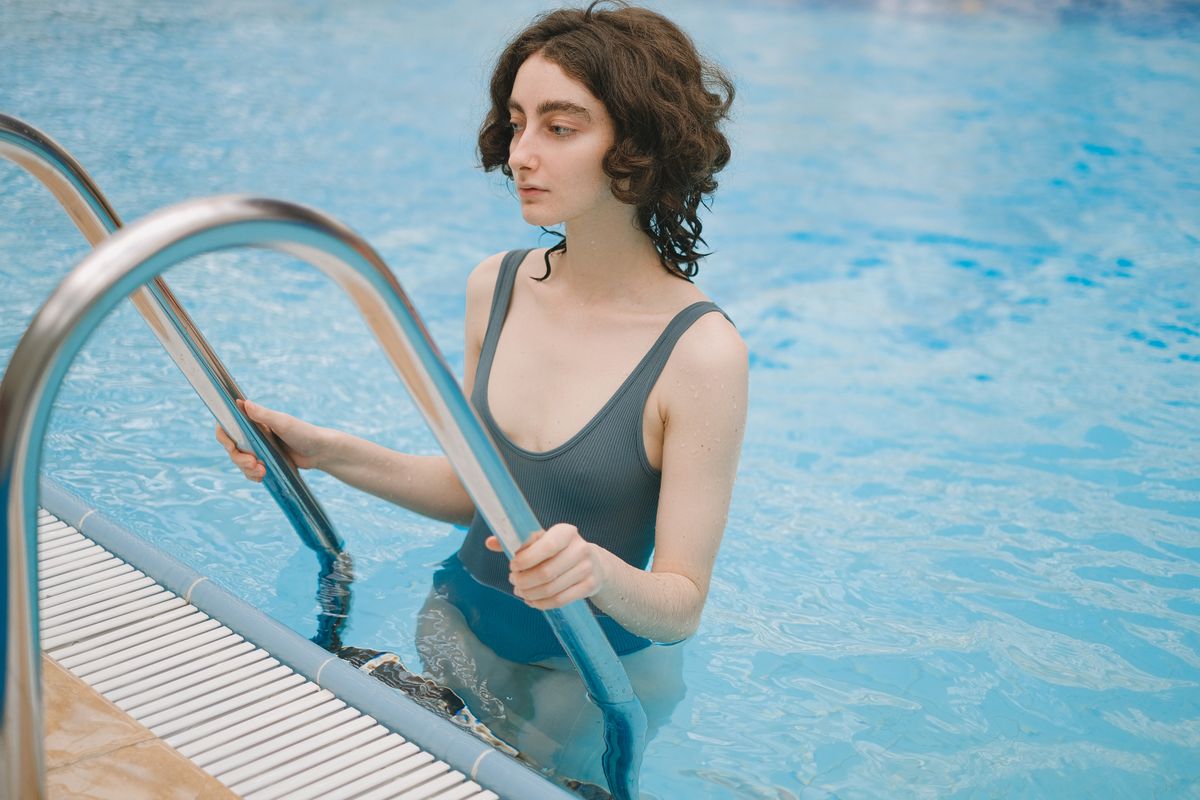 A young woman in a gray swimsuit holds onto poolside railings, standing in clear blue water.