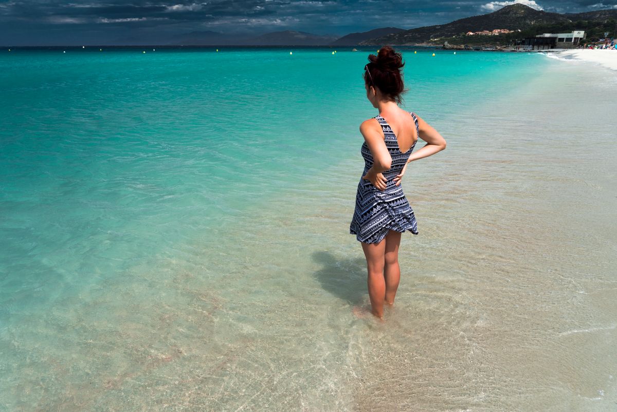 Woman standing on a beautiful sandy beach looking at turquoise sea under cloudy skies.