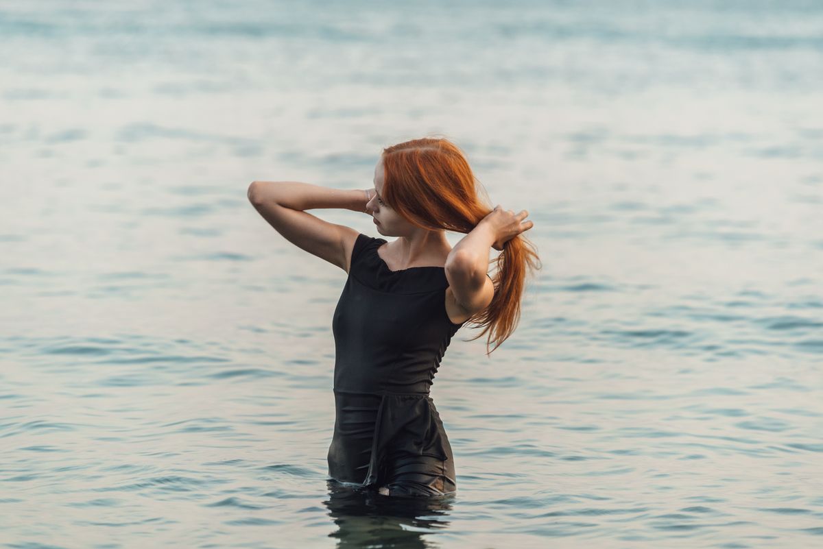 A woman stands in the water, holding her hair, surrounded by the calming ocean at sunset.