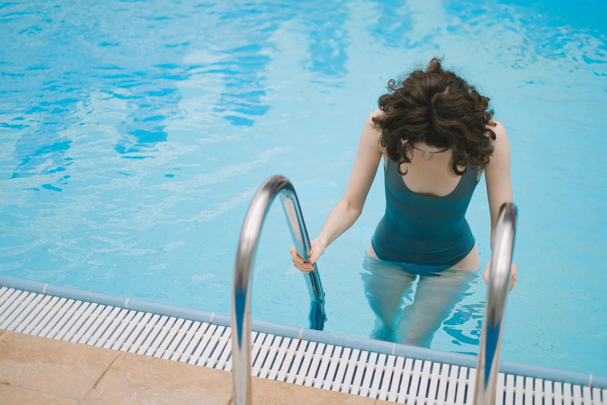 Young woman in a teal swimsuit using ladder to enter the pool on a sunny day.