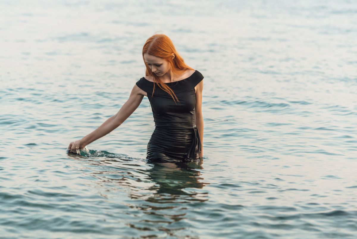 Red-haired woman in black dress wading through the sea in Гагра, showcasing serenity and elegance.