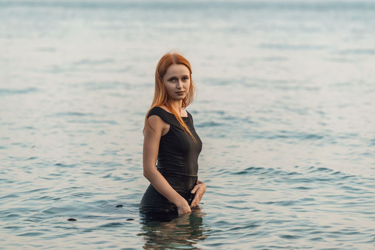A woman in a black dress gracefully standing in the calm sea during the day.