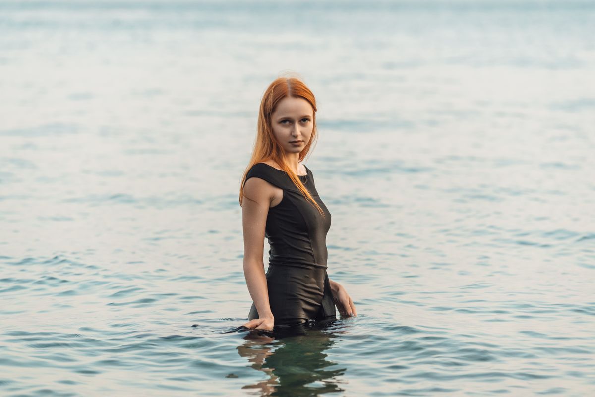 A young woman with long hair standing in the sea, wearing a black dress on a calm day.