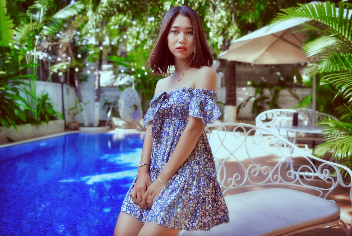 Woman in floral dress standing by a vibrant blue swimming pool, surrounded by lush greenery.
