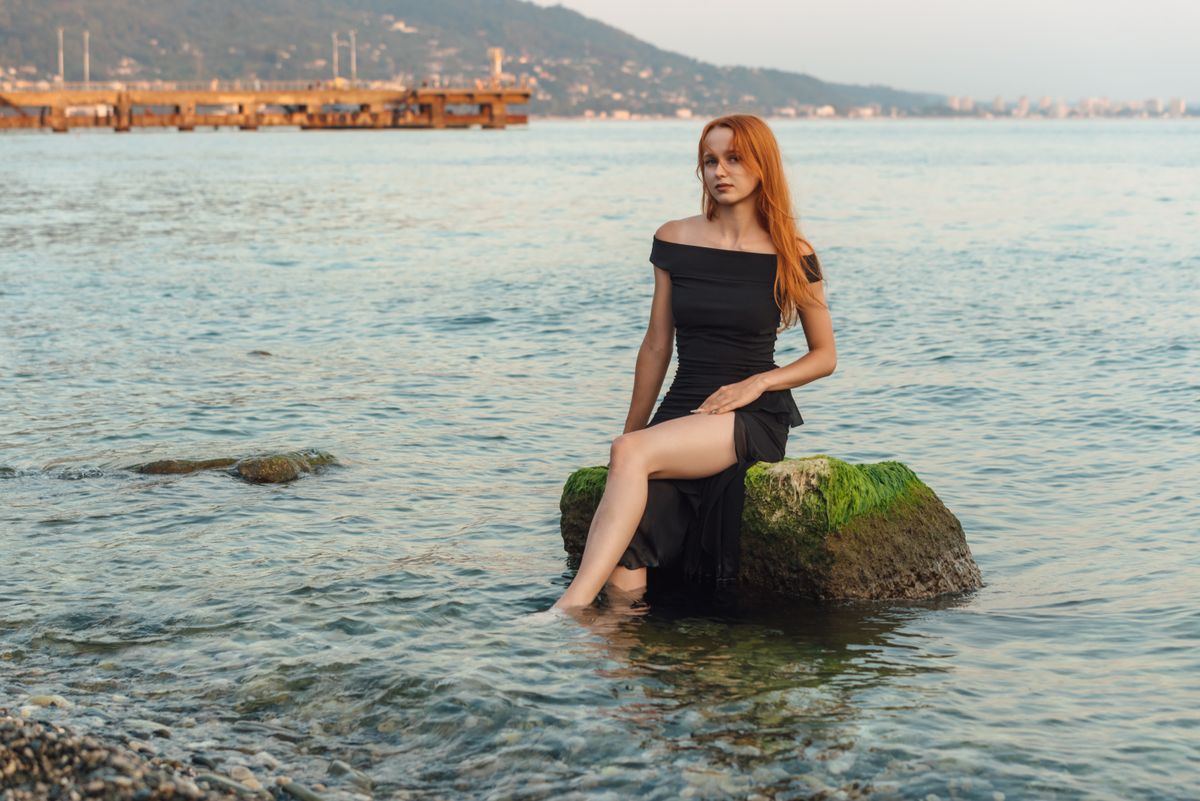A woman in a black dress sits gracefully on a moss-covered rock by the sea.