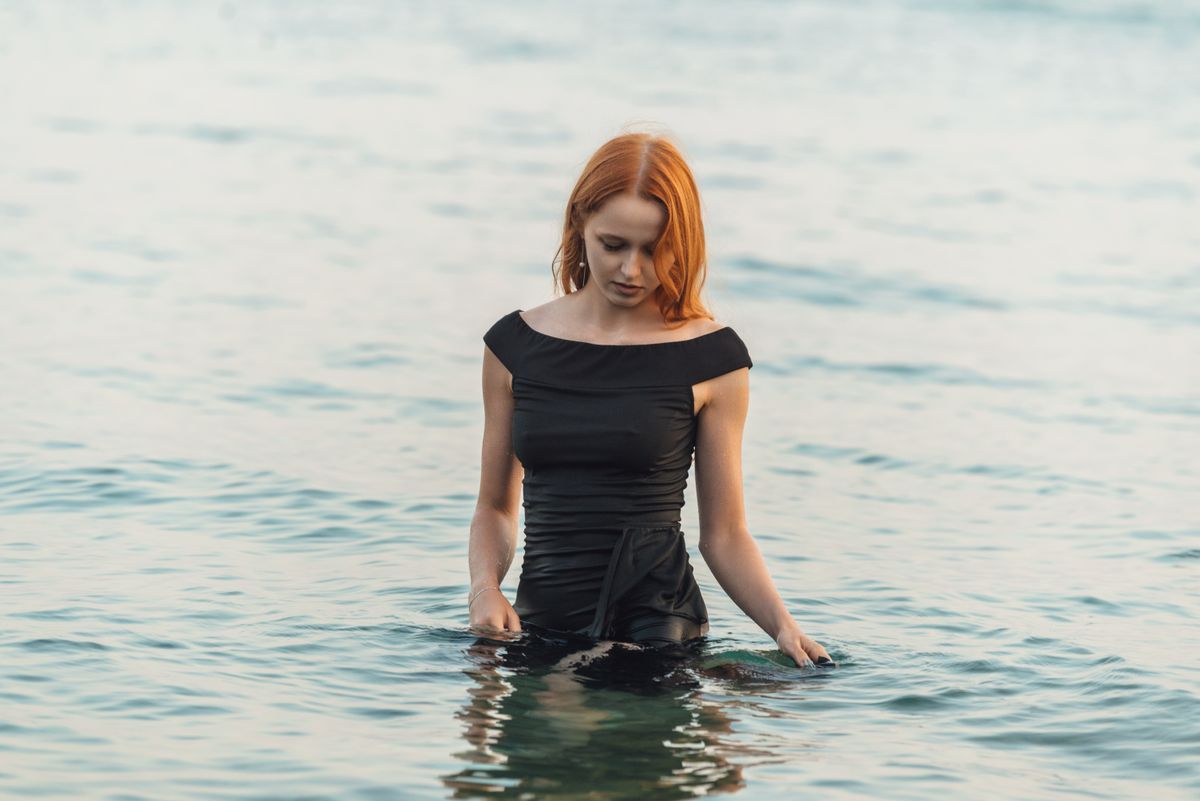 A woman with red hair wearing a black dress standing in a calm sea during the day.