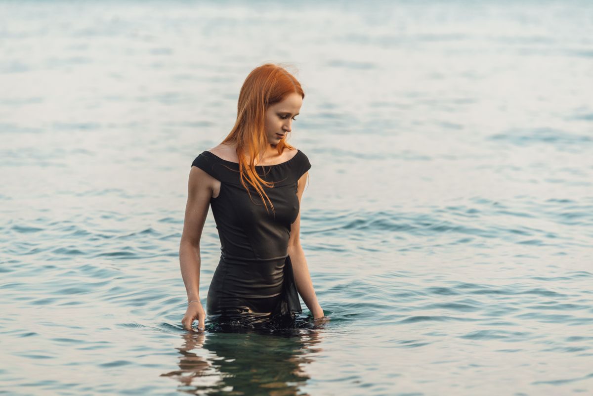 Thoughtful redhead woman in black dress standing in calm sea waters of Гагра.