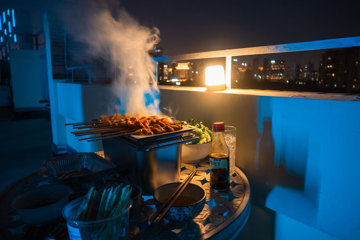 A cozy rooftop barbecue scene at night with skewers and city lights in the background.