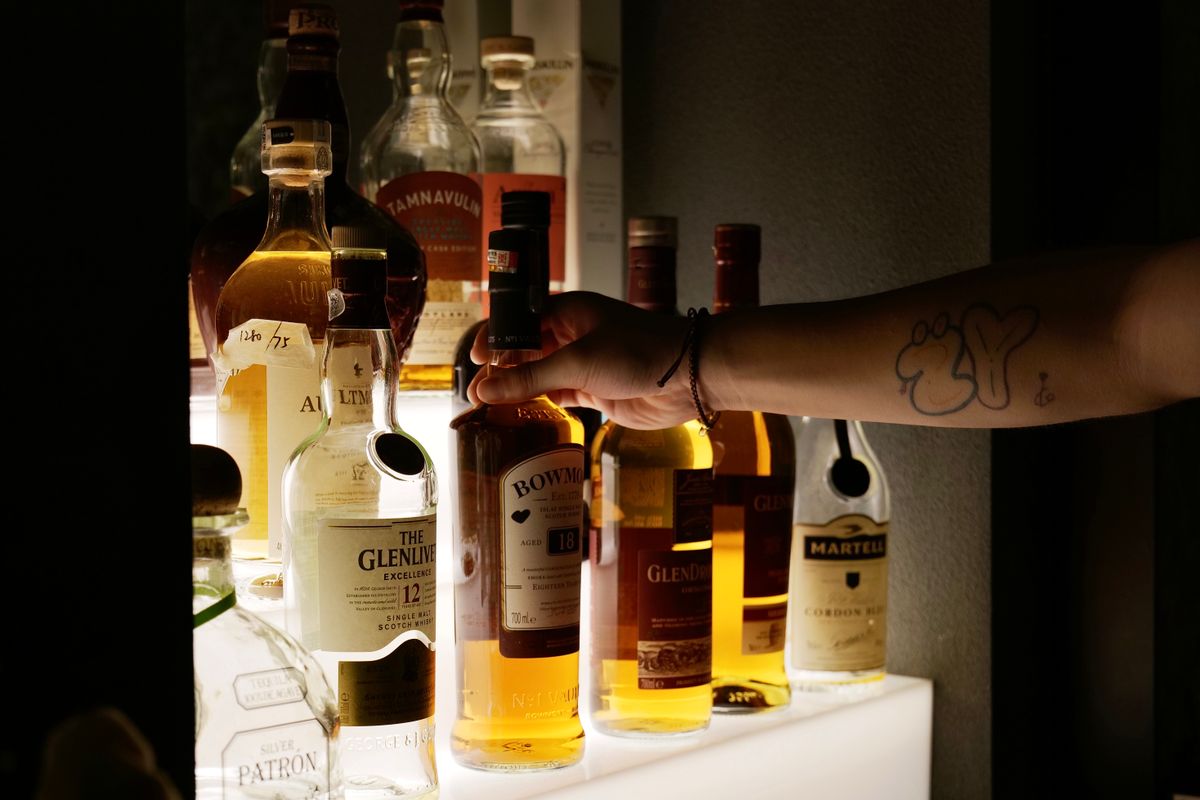 A tattooed hand selects a whiskey bottle from an illuminated shelf, showcasing various liquor options at a bar.