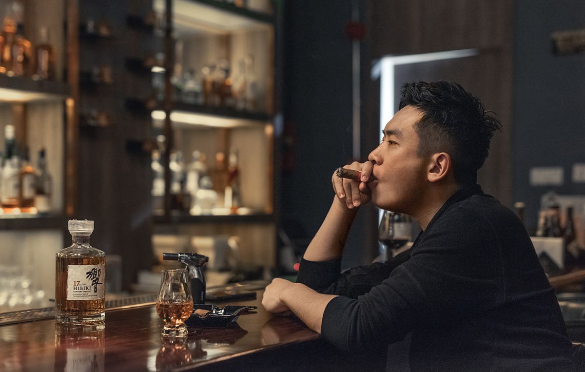 Side view of ethnic male in black shirt sitting alone at bar counter while smoking cigar and drinking Japanese whiskey against illuminated shelves with bottles of different alcohol