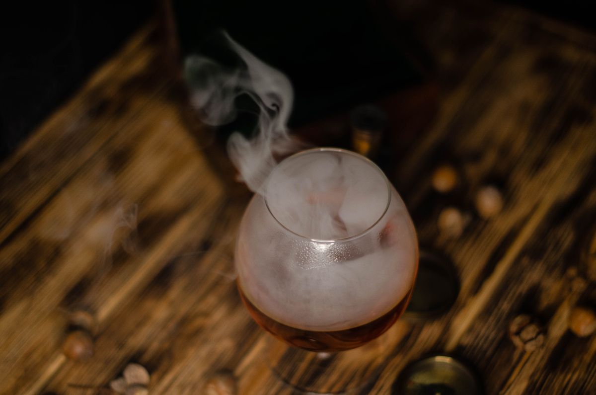 A smoky cocktail served in a glass on a rustic wooden table, surrounded by nuts and smoke.