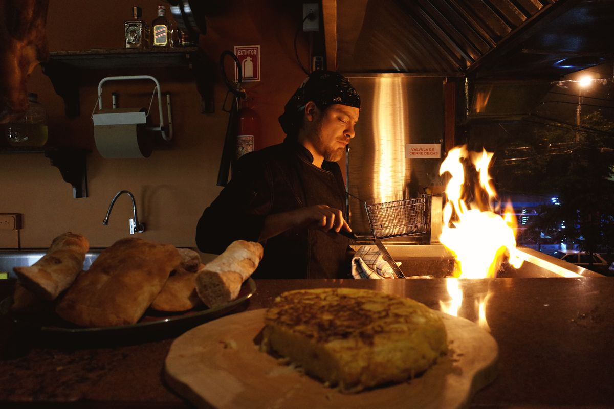 A chef expertly cooks at night beside a fire in a warm, dimly lit kitchen.