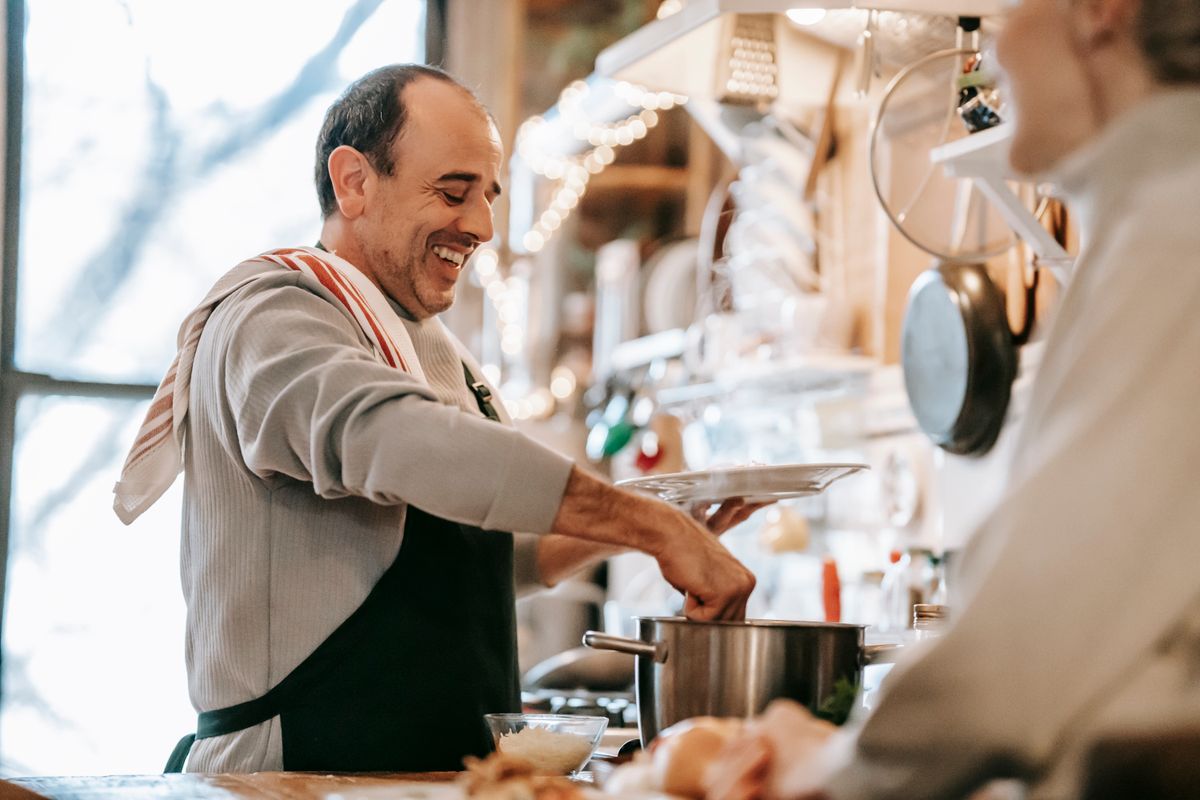 Side view of joyful adult ethnic male in casual clothes and apron standing at table in kitchen near anonymous wife while serving yummy homemade food during dinner