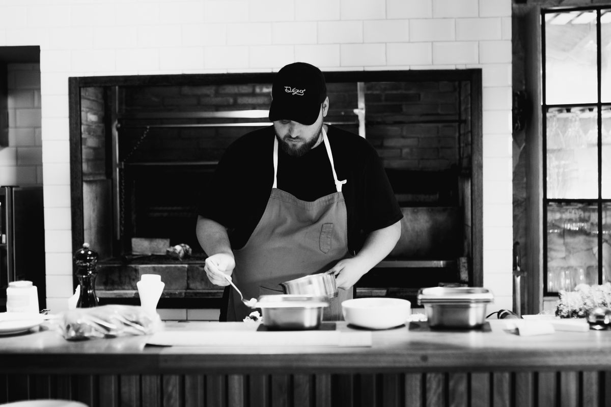 A chef wearing an apron prepares a meal in a rustic kitchen. Black and white photography.
