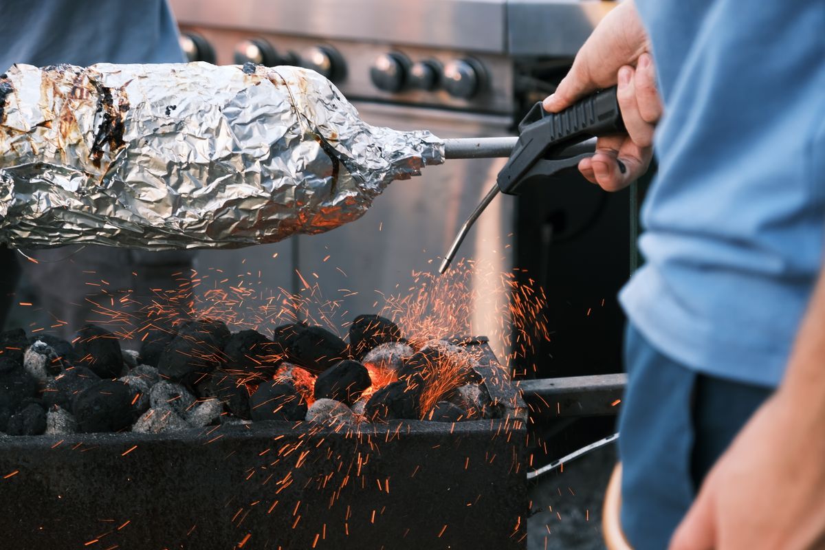 Barbecue cooking with foil-wrapped food over charcoal flames showing flying sparks and intense heat.