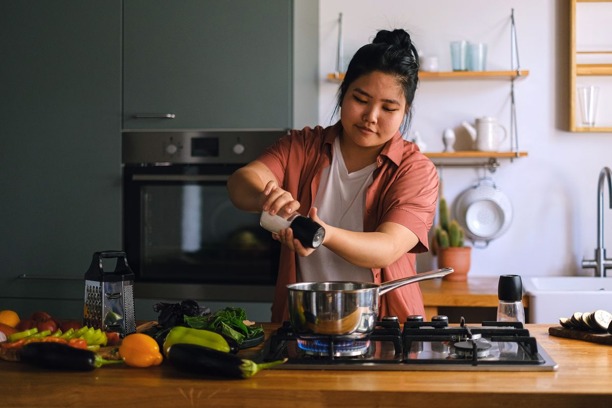 A home cook seasoning a dish on a stove surrounded by fresh vegetables in a modern kitchen.