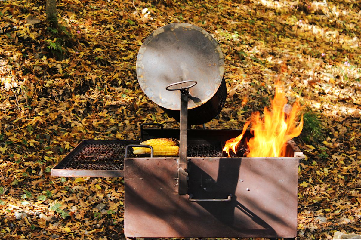 Rustic grill with fire and corn in autumn leaves setting, perfect for outdoor cooking vibes.