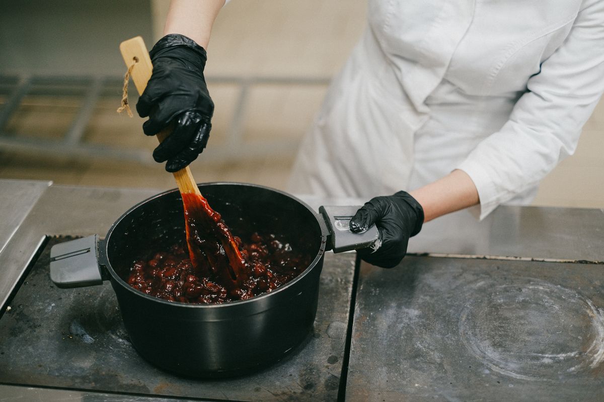 Chef stirring sauce in a pot with black gloves, showcasing food preparation techniques.
