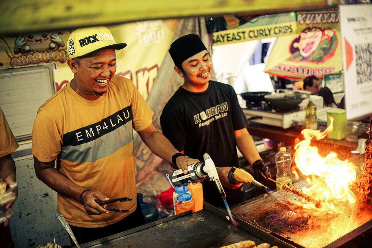 Cheerful street food vendors using a torch to cook skewers at a vibrant market.